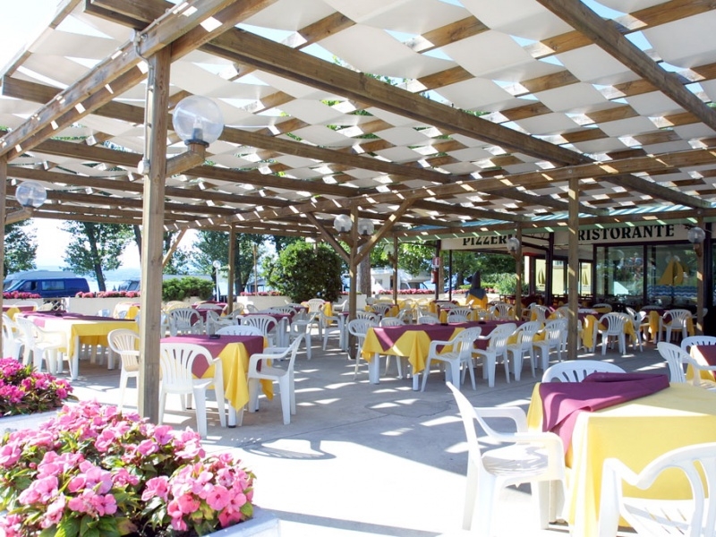 Tables and chairs arranged under a wooden pergola, surrounded by flowers and greenery at San Francesco