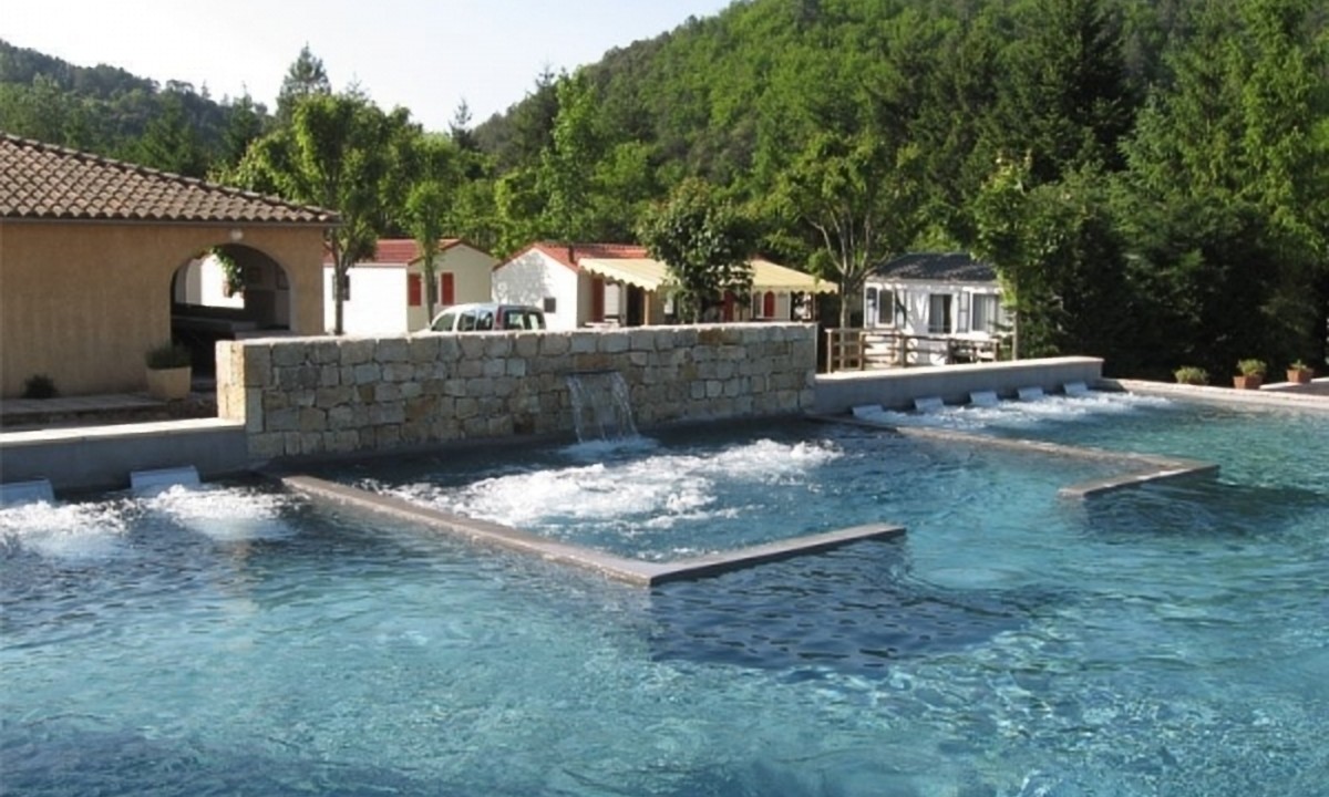 At Les Ranchisses, there is a swimming pool equipped with water jets and a stone wall feature, encircled by lush trees and small structures.