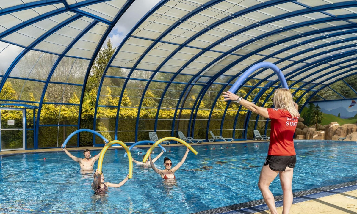 Instructor leads a fitness class using pool noodles in an indoor swimming pool with a glass roof at La Ribeyre