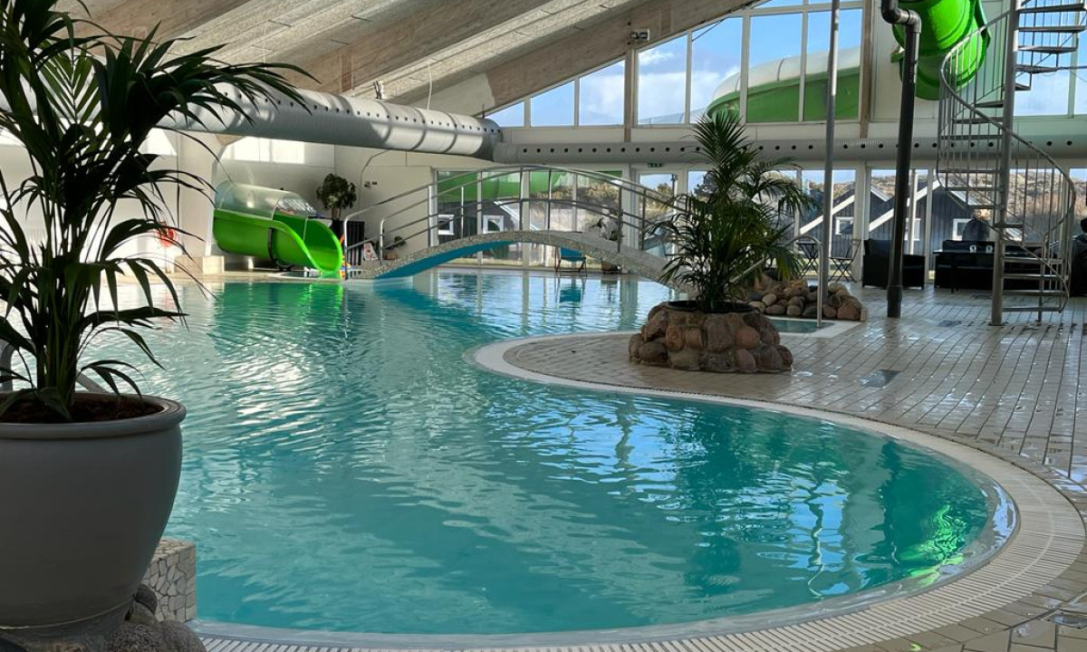At Dancamps Nordsø, there is an indoor swimming pool featuring a twisting waterslide, encircled by potted greenery and expansive windows.