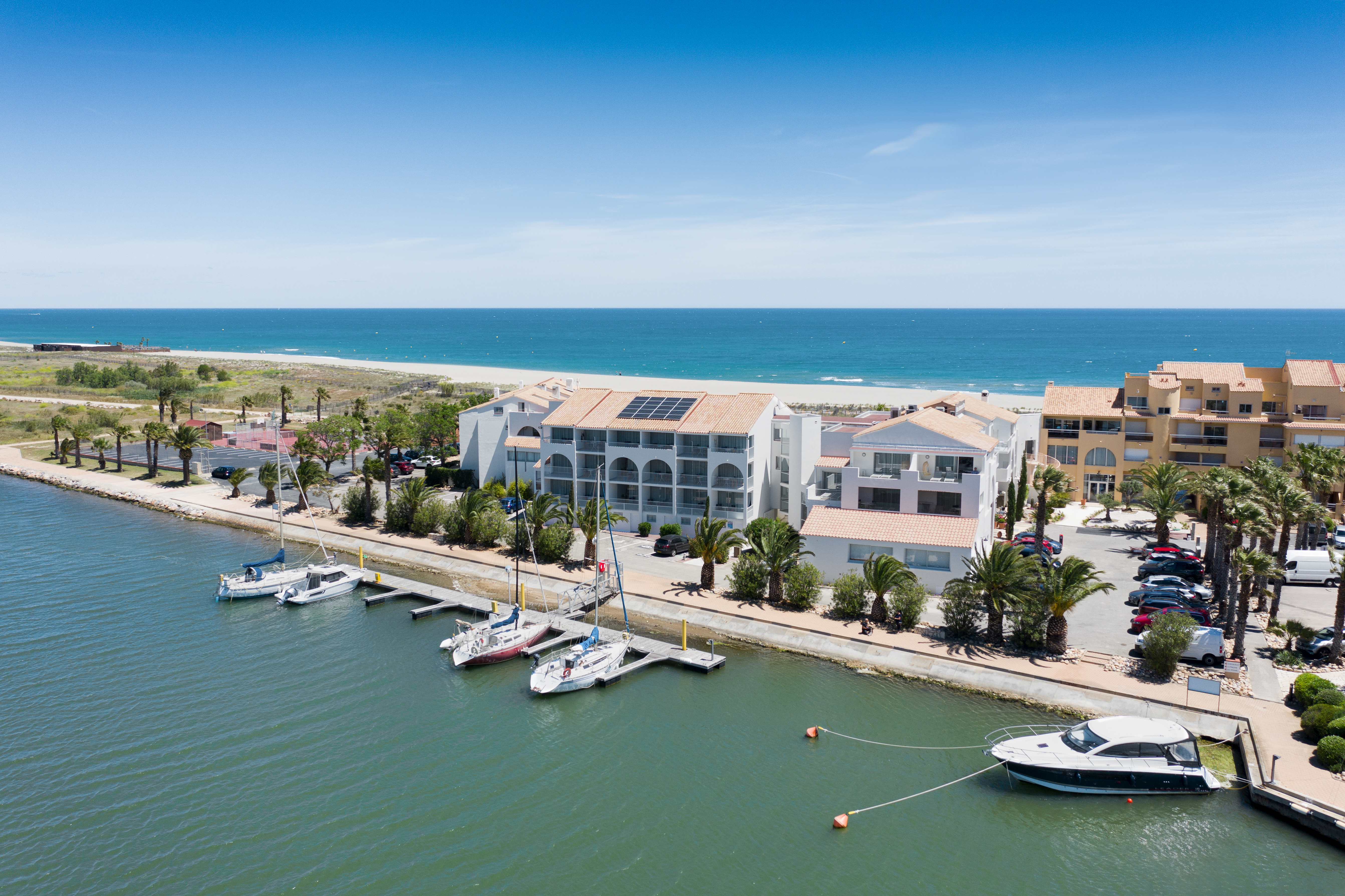 Several yachts moored at a marina; adjacent buildings with palm trees; distant ocean and beach under clear blue sky at Les Bulles de Mer