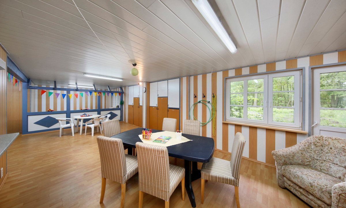 At KNAUS Campingpark Wingst, there is a dining table accompanied by striped chairs in a vibrant, adorned room featuring windows that provide a view of a forest.