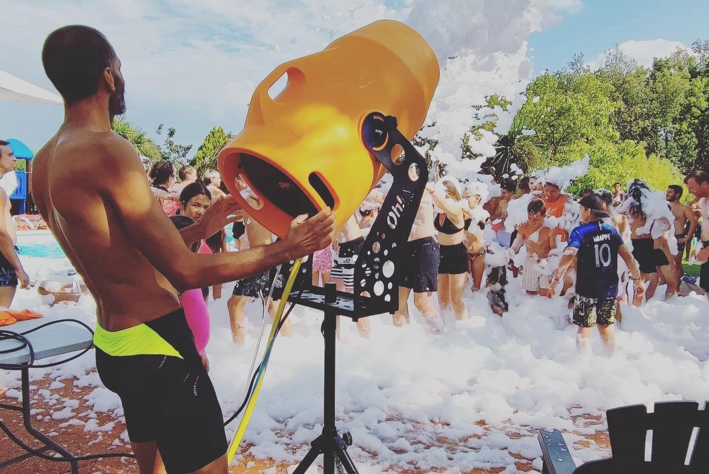 At Domaine du Koukano, a man operates a foam machine while joyful individuals are enveloped in foam at a poolside celebration.