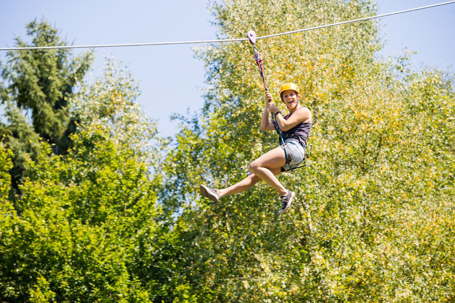 At Parc La Clusure, an individual is zip-lining while wearing a yellow helmet amidst a wooded area, with vibrant green vegetation in the backdrop.