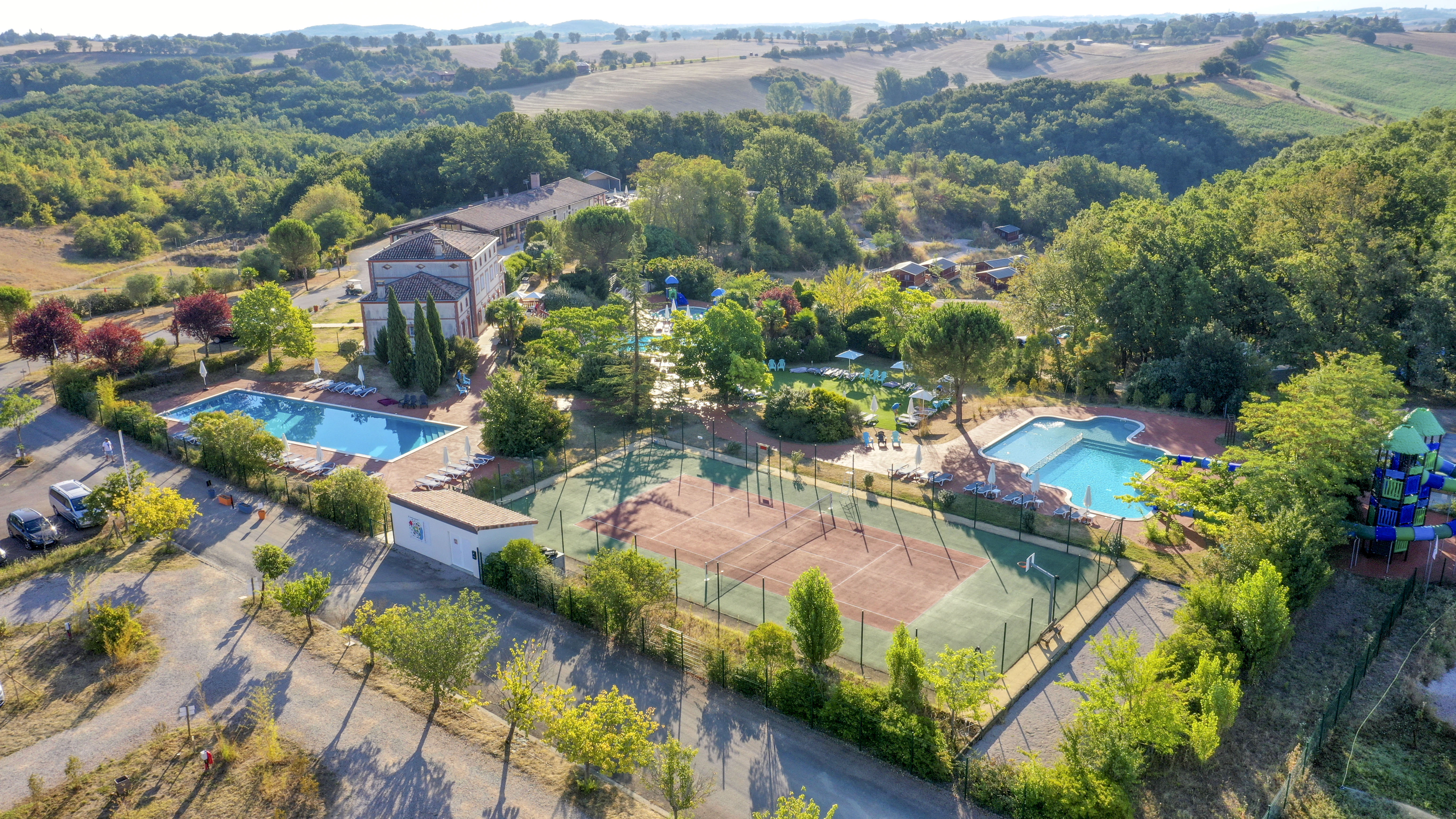 At Domaine du Koukano, a swimming pool is situated next to a building and tennis courts, enveloped by vibrant greenery and a remote countryside view.