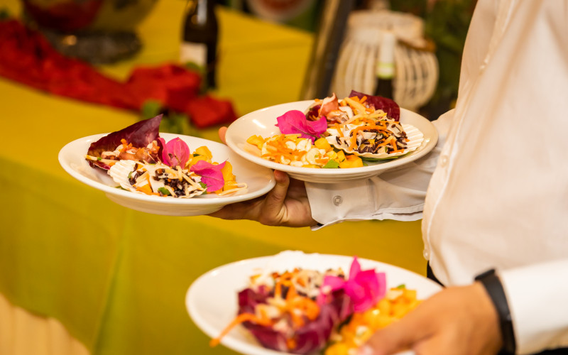Server carrying three colorful salad plates, garnished with flowers, at a festive indoor dining area at Triscina Mare