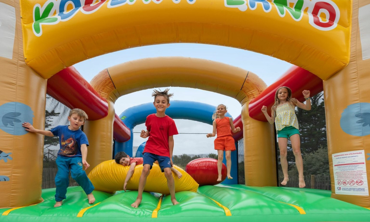 Children joyfully jumping on a colorful inflatable bouncer, surrounded by trees, under a cloudy sky at Maguide