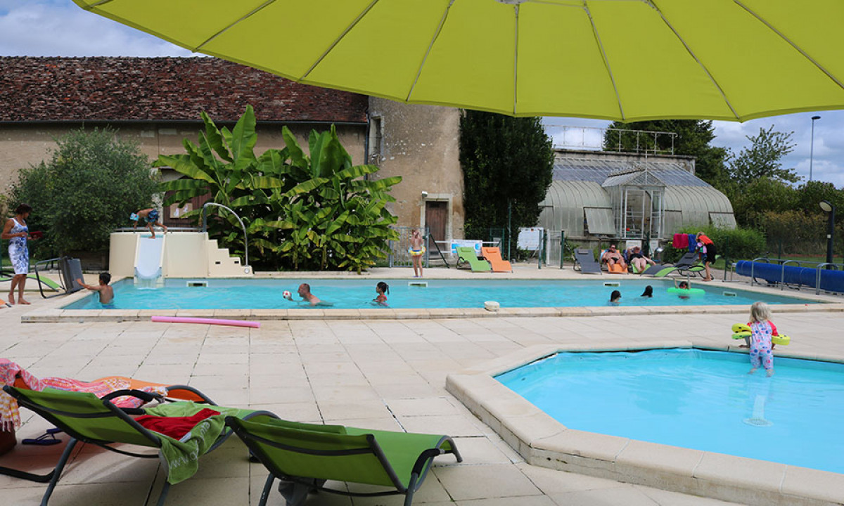 At Le Petit Trianon, a swimming pool features children playing, encircled by loungers and lush greenery beneath a large yellow umbrella.