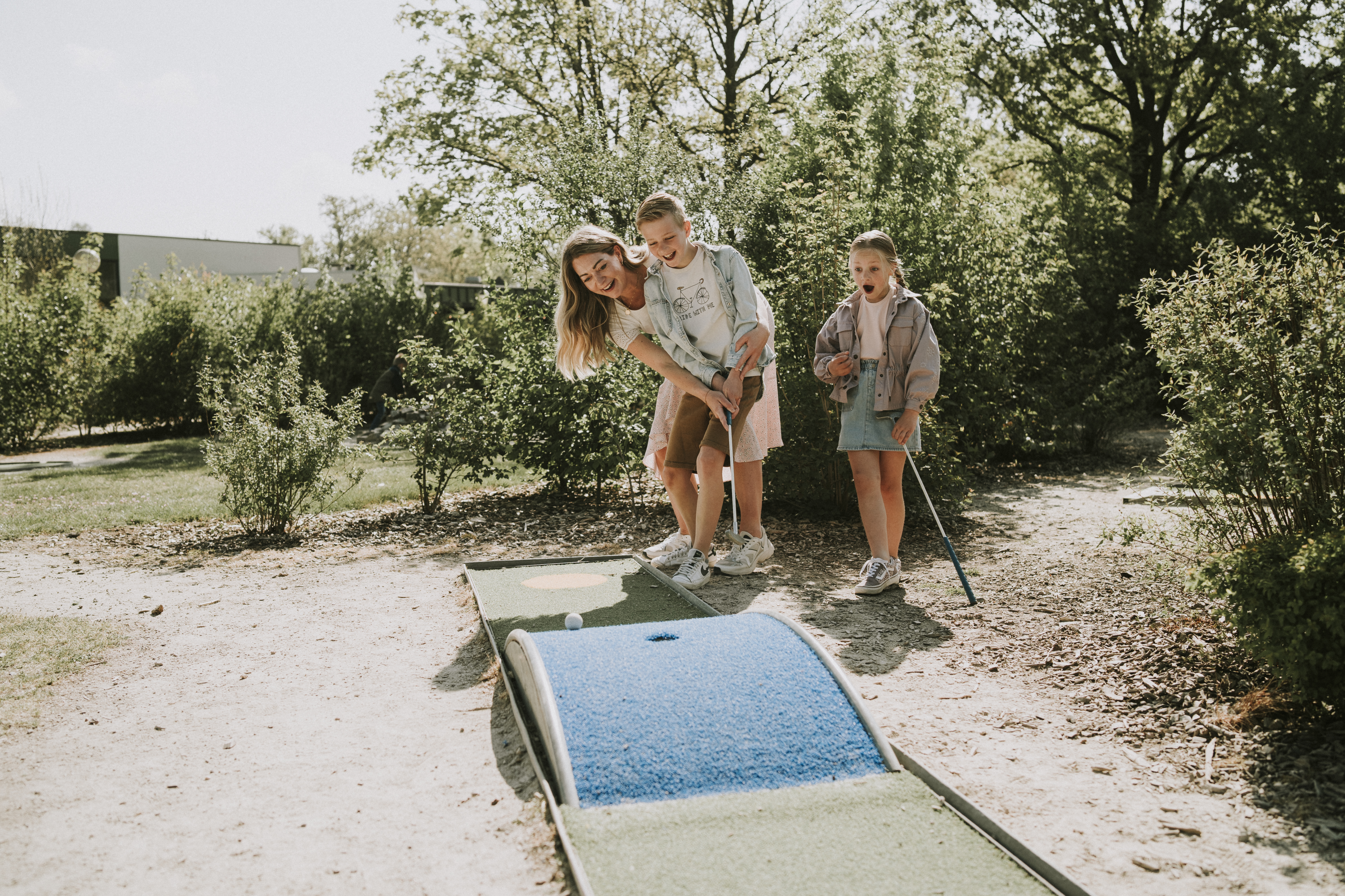 At Vakantiepark Dierenbos, an adult helps a child play mini-golf while another child observes with enthusiasm. Lush greenery encircles the course area.