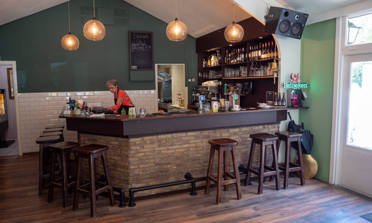 Bartender cleaning bar counter in cozy pub with wooden stools, hanging lights, and shelves stocked with bottles at de Wrange