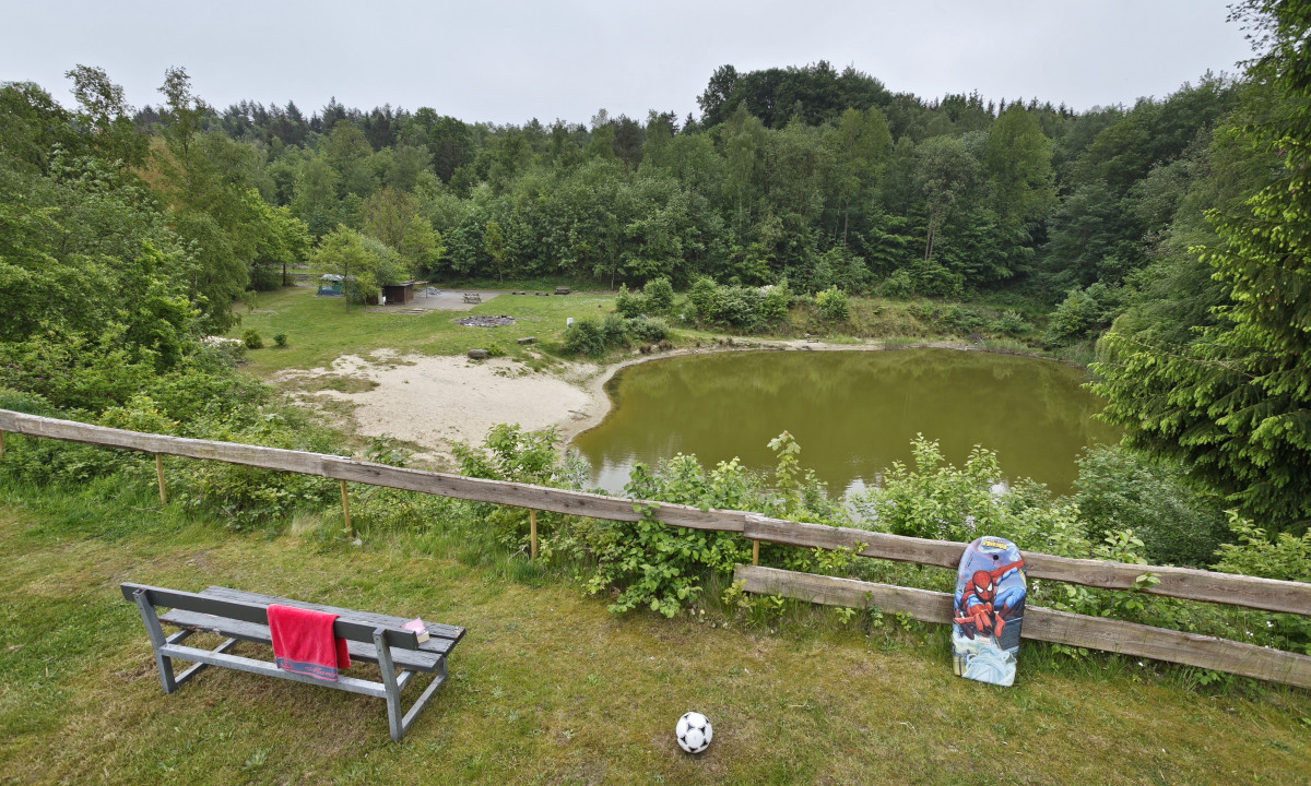 At KNAUS Campingpark Wingst, a wooden seat provides a view of a small, verdant lake, surrounded by trees and a sandy area.