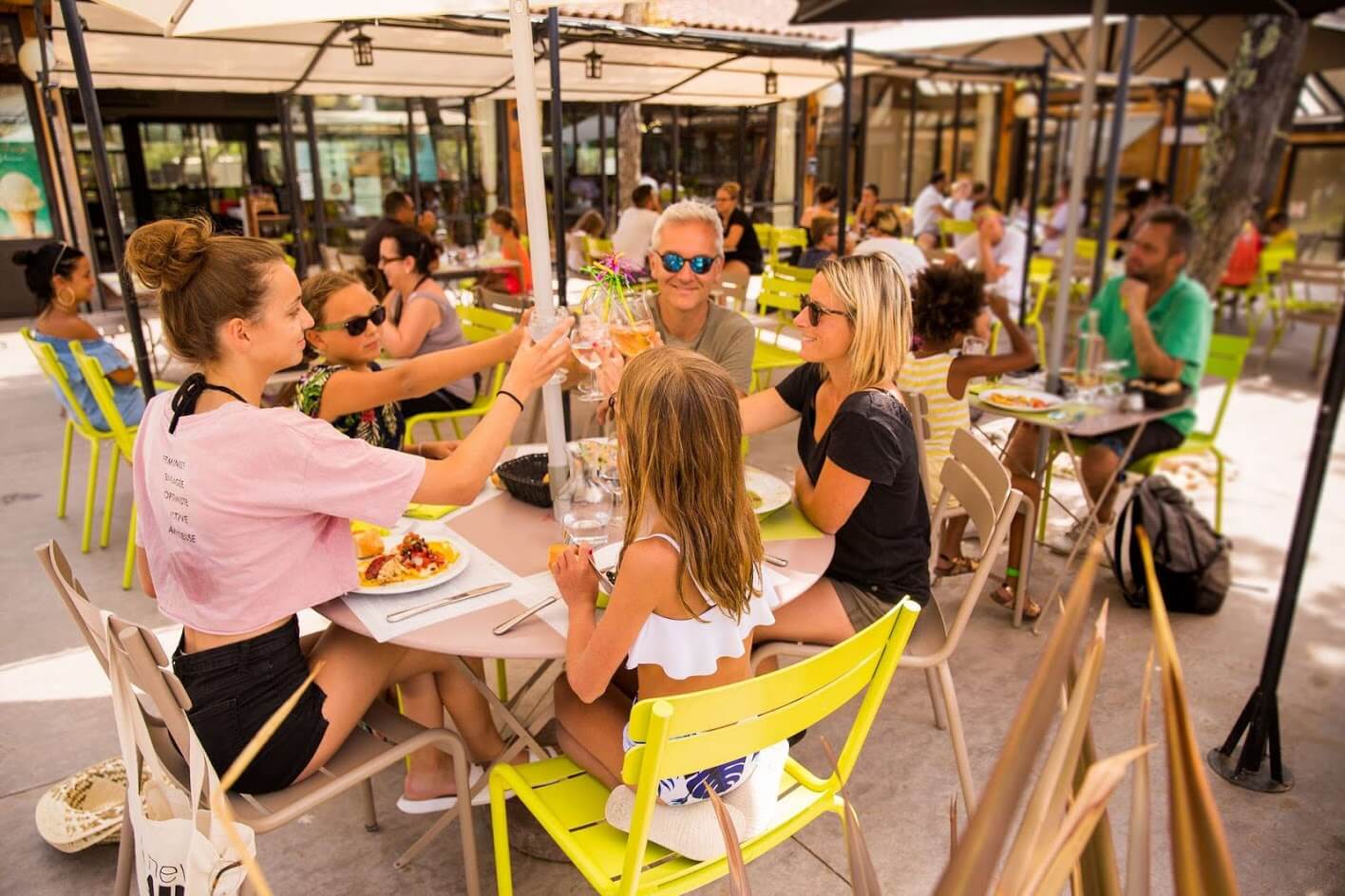 At Le Ranc Davaine, individuals are savoring a meal while raising their glasses at an outdoor café featuring yellow chairs.