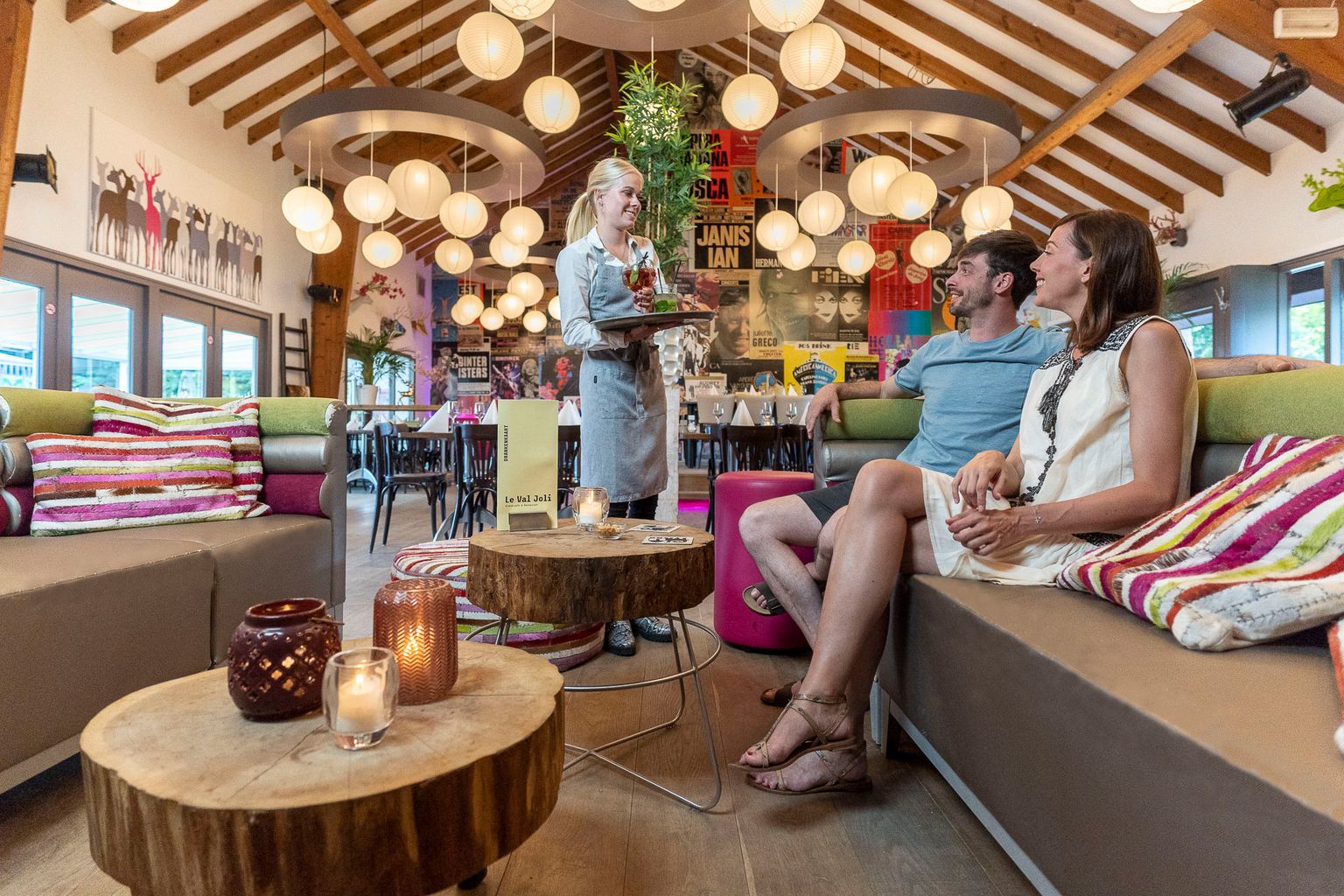 At Parc La Clusure, a server is offering a dessert to a seated couple in a warm, lantern-lit café featuring wooden beams and vibrant decor.