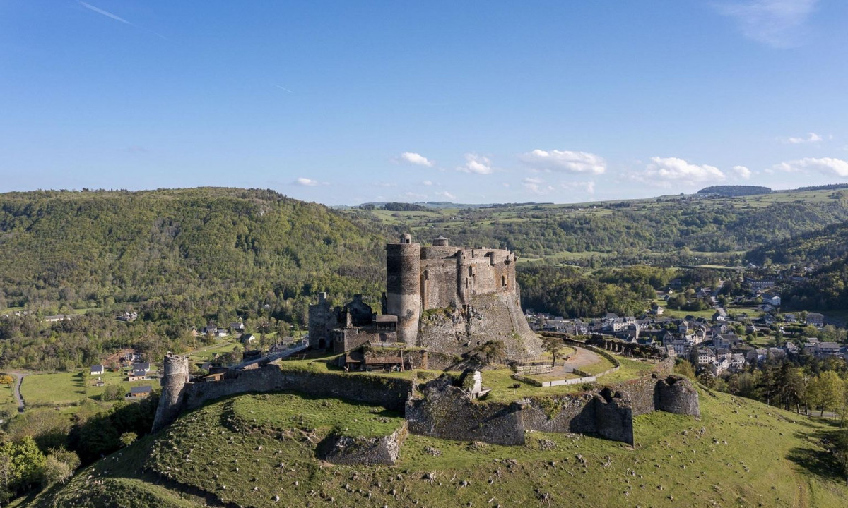 Ancient stone castle overlooks green valley, surrounded by scattered houses and distant wooded hills at La Ribeyre