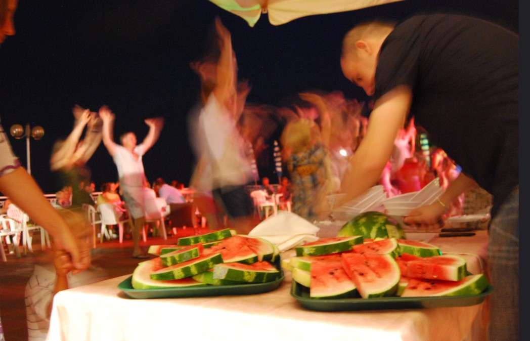 Slices of watermelon on a table being served, people dancing in the background at Sant' Anna Residence
