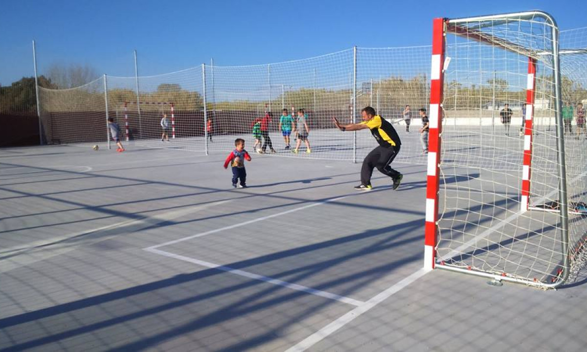 A child runs toward an adult goalkeeper on an outdoor soccer court with multiple players at Resort Els Pins