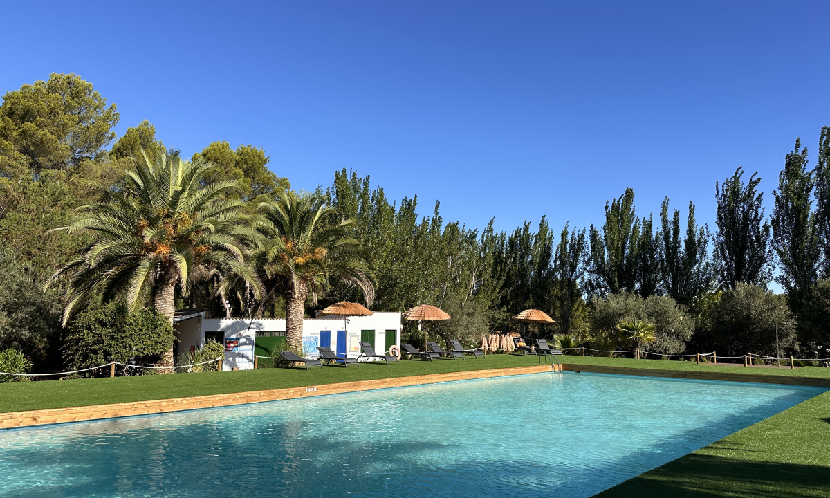 At Taïga Lake Caspe, a swimming pool glimmers beneath clear blue skies, encircled by sunbeds, palm trees, and vibrant foliage.