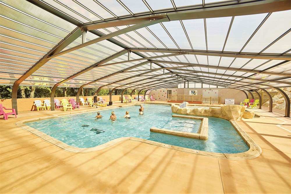 Covered swimming pool with several people swimming, surrounded by lounging chairs and natural rock formations at Les Albères