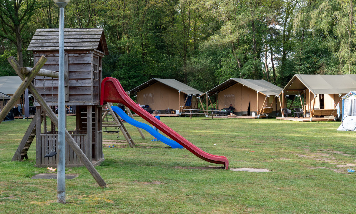 Playground structure with slide beside a lamppost, in front of tents set up in a wooded campsite area at de Wrange