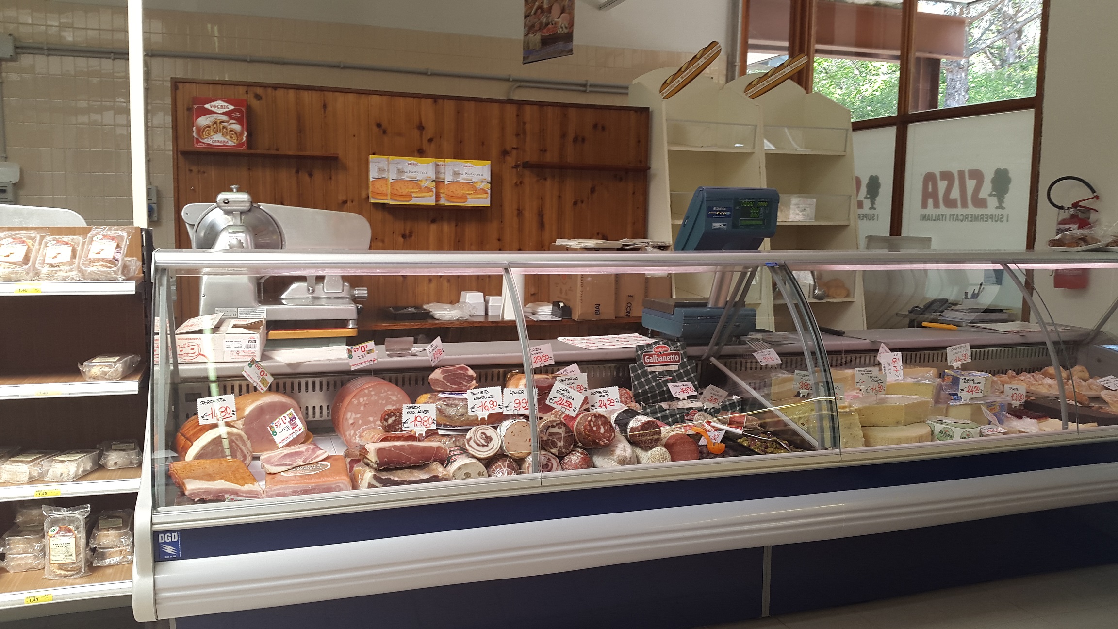 Deli counter displaying various meats and cheeses with slicer; wooden panel wall in background, store shelving on left at Belvedere Pineta