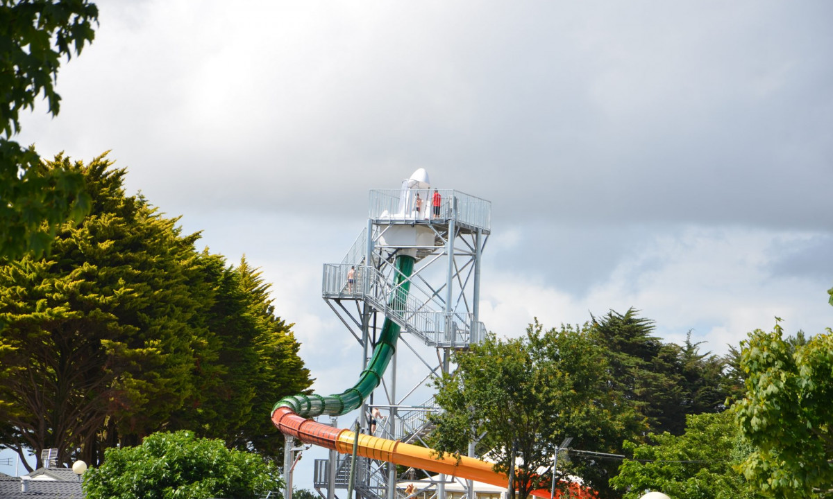 At Le Domaine de Léveno, a waterslide tower rises amidst a lush, tree-filled campground under overcast skies.