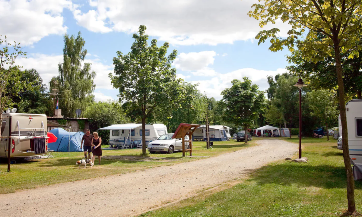 At KNAUS Campingpark Lübben, a pair strolls with their canine along a dirt trail encircled by caravans and tents among the trees.