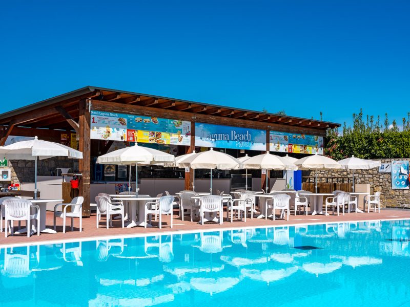Outdoor cafe with white umbrellas and plastic chairs in front of a swimming pool under clear blue skies at Residence Eden