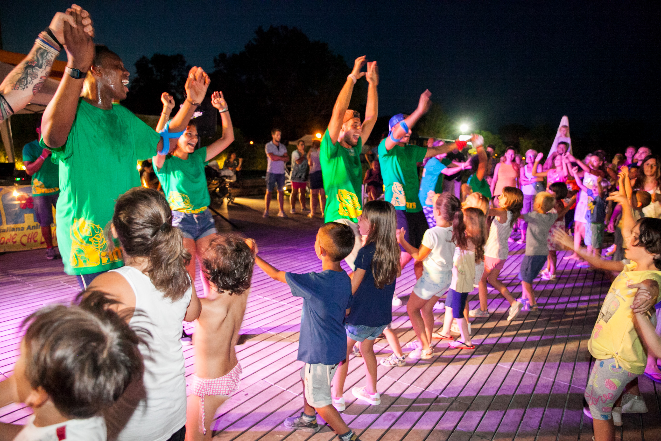 Adults lead children in a lively dance under colorful lights on a wooden deck at Pineta Sul Mare Camping Village