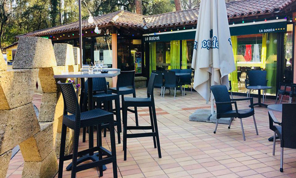 At La Sousta at the front, elevated tables and stools are positioned on a tiled terrace in front of a charming café adorned with string lights, encircled by trees.