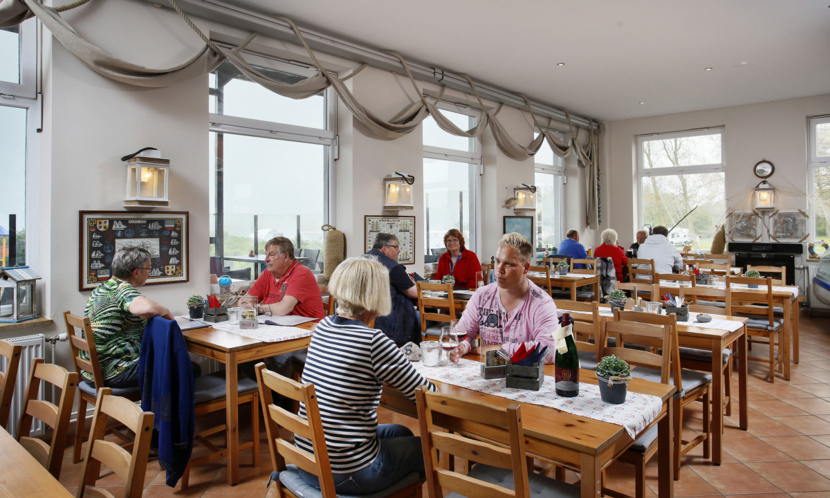 At KNAUS Campingpark Rügen, individuals are enjoying meals at wooden tables in a vibrant, maritime-themed eatery featuring expansive windows.