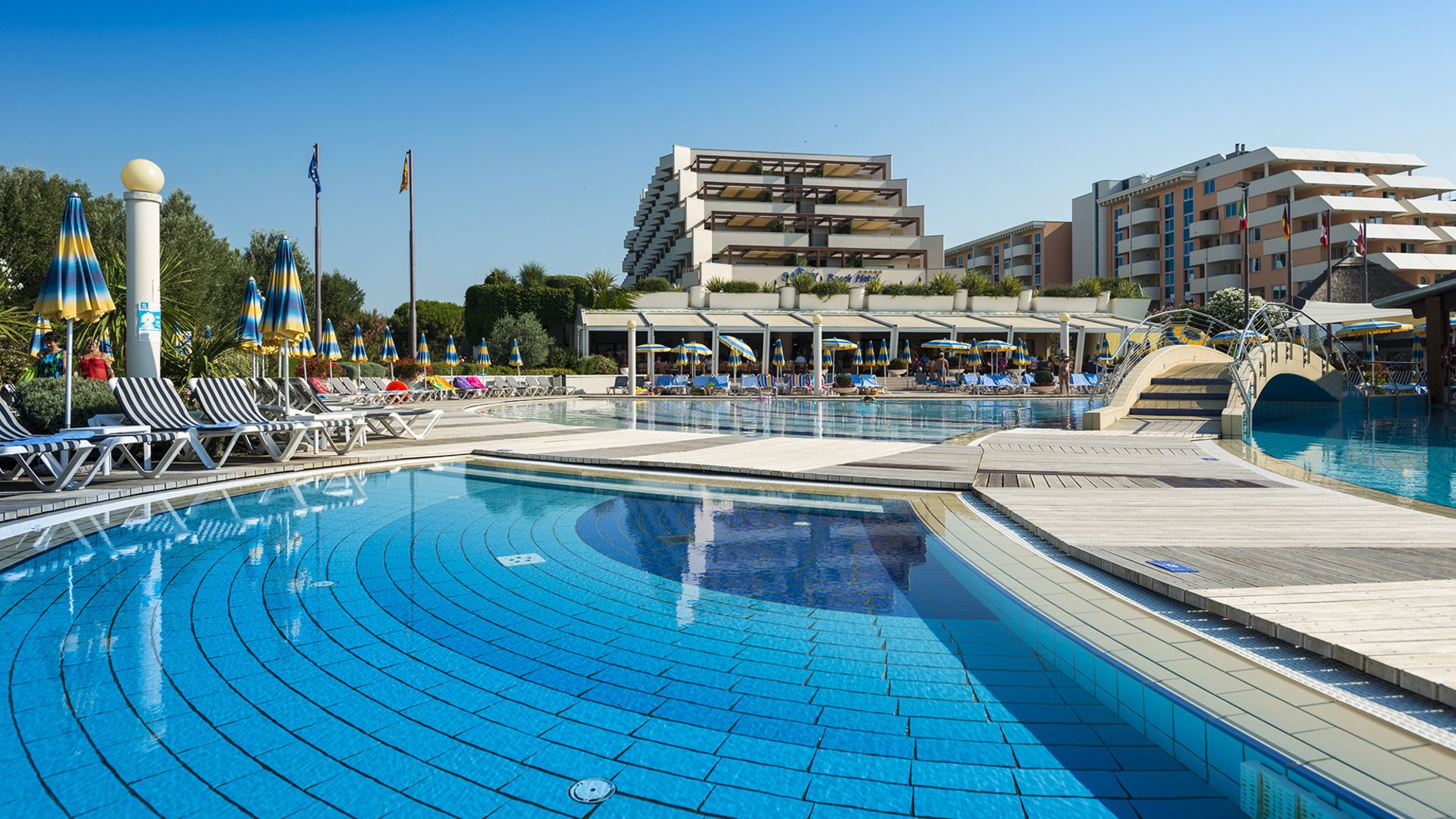 A clear pool with bridge and lounge chairs, surrounded by buildings and umbrellas under a sunny sky at Savoy Beach Hotel & Thermal Spa