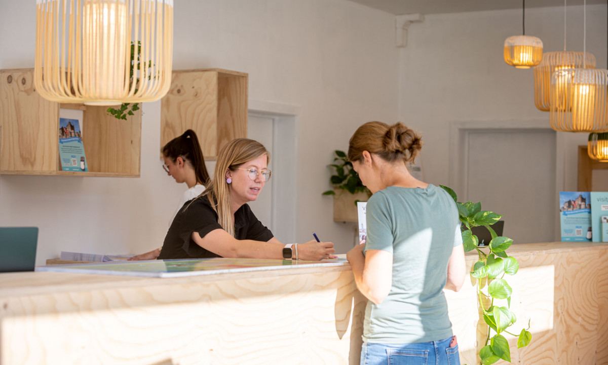 At Urban Gardens Gent, two women are engaged at a wooden reception counter, one jotting down notes while the other is performing tasks, all set against a backdrop of contemporary decor.