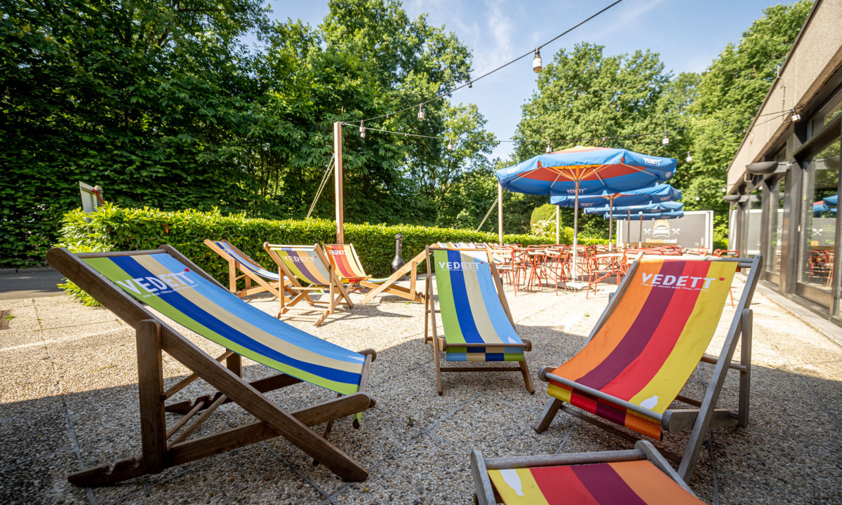 At Urban Gardens Gent, deck chairs are positioned in a sunlit patio space, encircled by lush greenery and outdoor umbrellas and tables.
