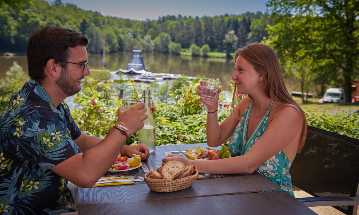 At Coucou, two individuals are savoring a meal and beverages at an al fresco table, gazing at a tranquil lake encircled by trees.