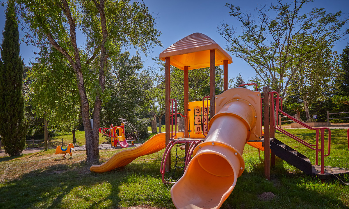 At Le Chêne Vert, there is a play area featuring slides and a tube, located in a sunny park with trees and grassy surroundings.