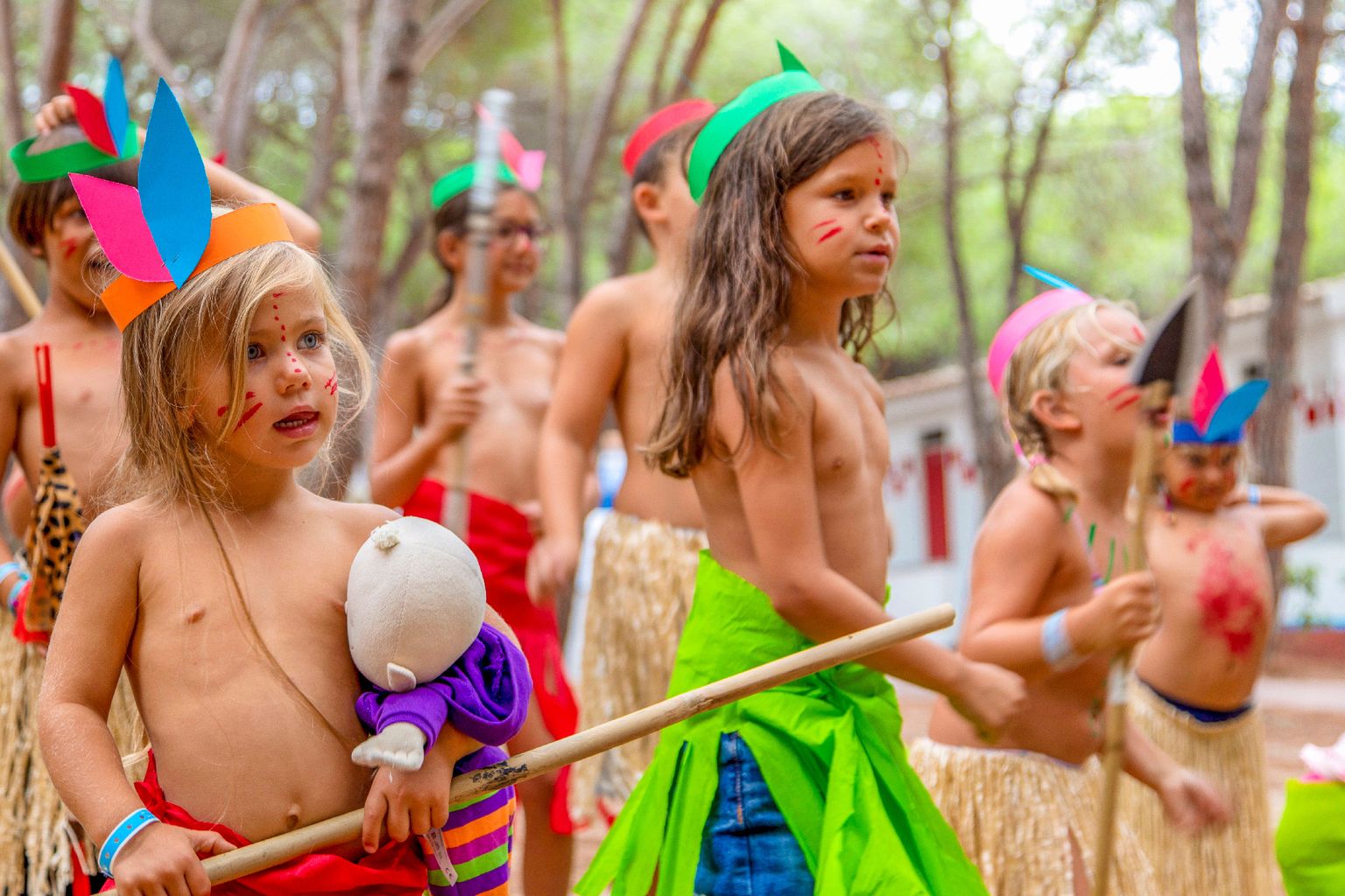 Children dressed in colorful costumes and face paint, holding sticks, parading outdoors among trees at Baia Blu La Tortuga