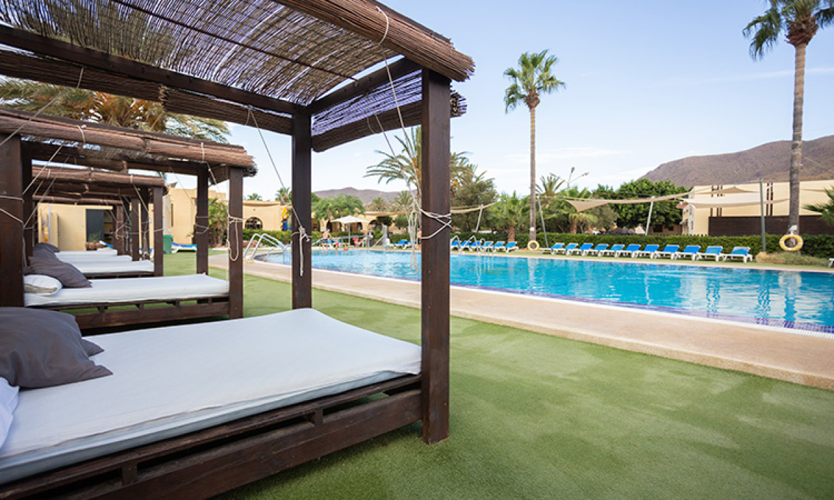 At Camping Los Escullos, wooden recliners with cushions are positioned next to a swimming pool surrounded by palm trees and mountains.