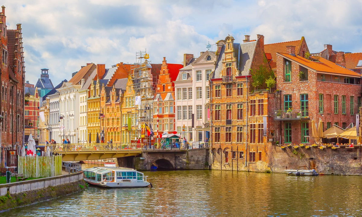At Urban Gardens Gent, a vessel is moored on a waterway next to vibrant, historic European structures beneath an overcast sky, with a bridge visible in the distance.
