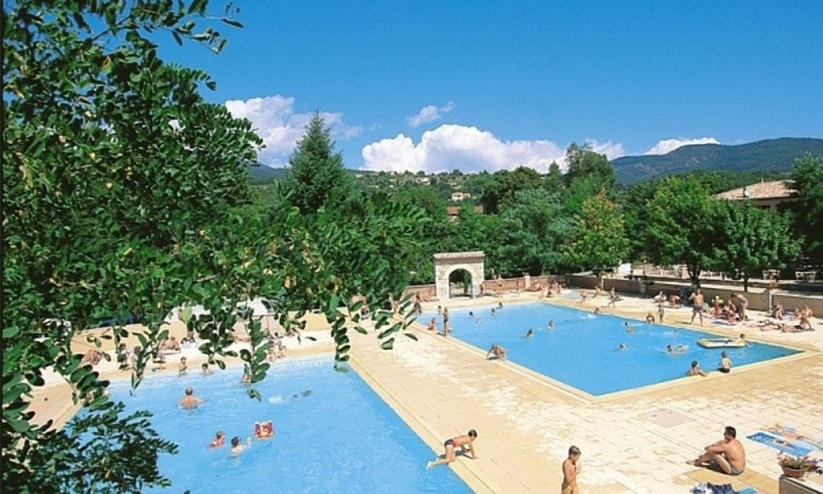 At Les Ranchisses, numerous individuals swim and unwind at a spacious outdoor swimming pool encircled by trees and mountains.