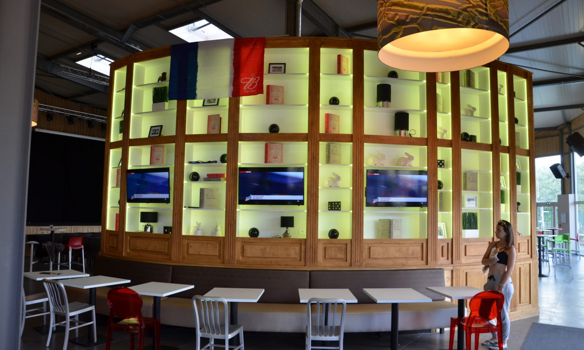 At Le Domaine de Léveno, there is a shelf-lined wall featuring televisions and a French flag, encircled by tables and chairs in the open dining space.
