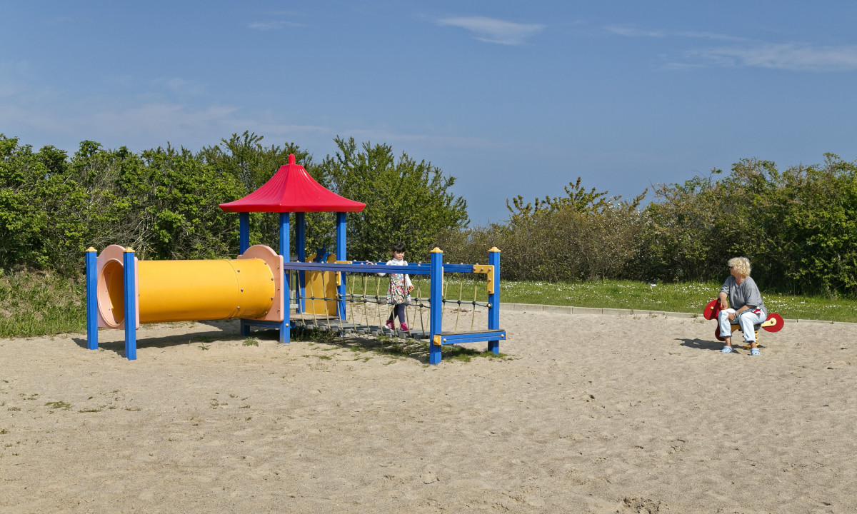 At KNAUS Campingpark Rügen, a play area features vibrant equipment and a child traversing a bridge; an adult is seated on a spring rider in a sandy zone, encircled by lush greenery.