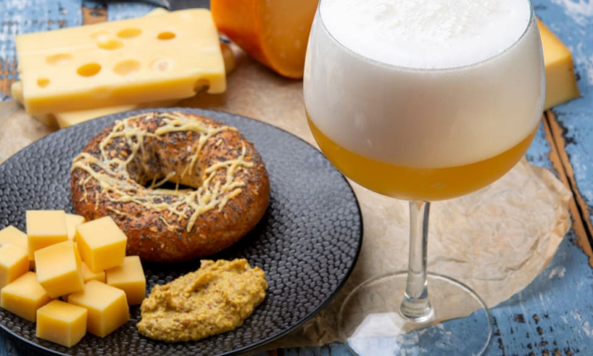 At Urban Gardens Gent, a bagel topped with cheese and mustard rests on a plate beside a frothy beer on a weathered wooden table.