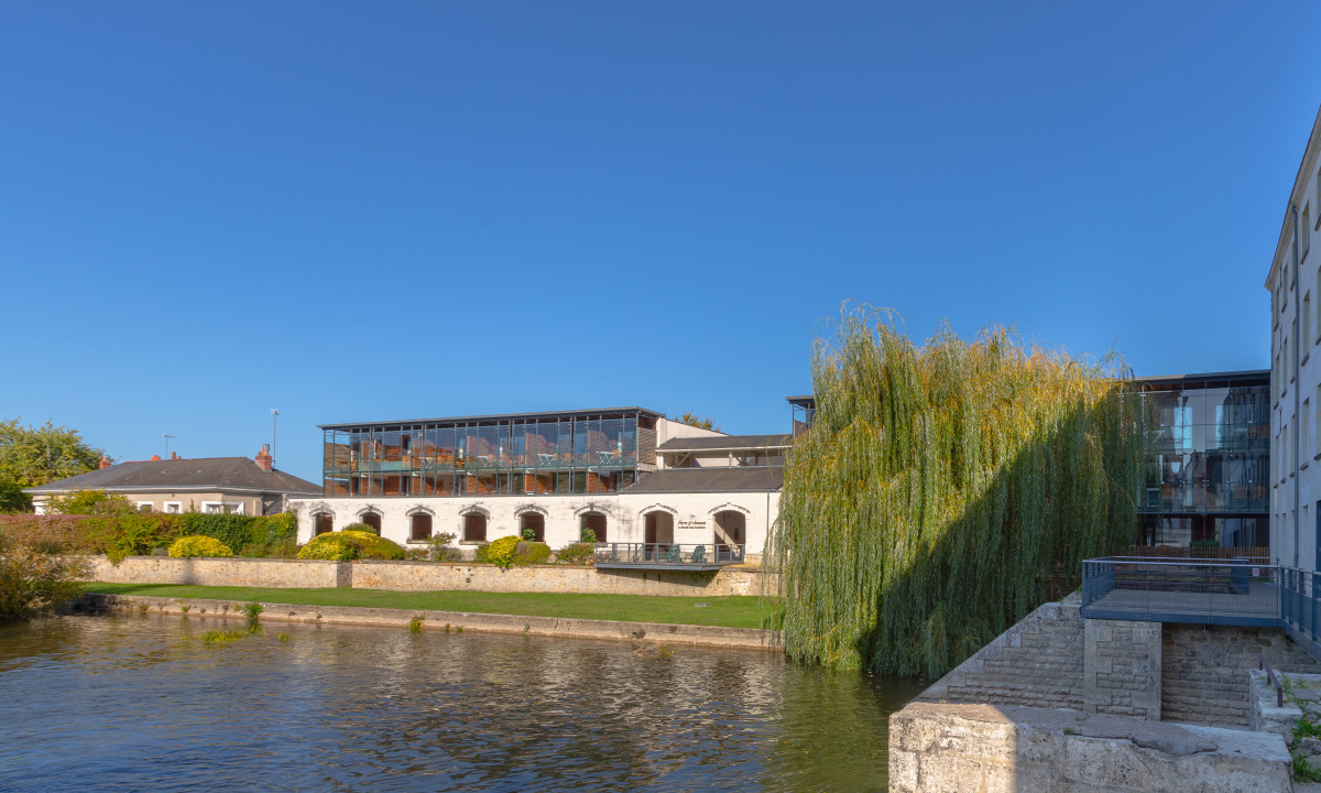 Building with large glass windows, situated by a riverside, surrounded by greenery and a clear blue sky at Le Moulin des Cordeliers