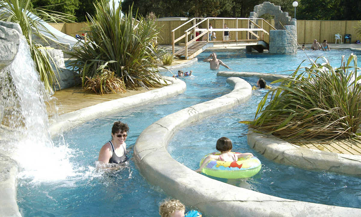 At Le Domaine de Léveno, individuals are swimming and drifting in a meandering, tropical-inspired pool featuring cascades and walkways.