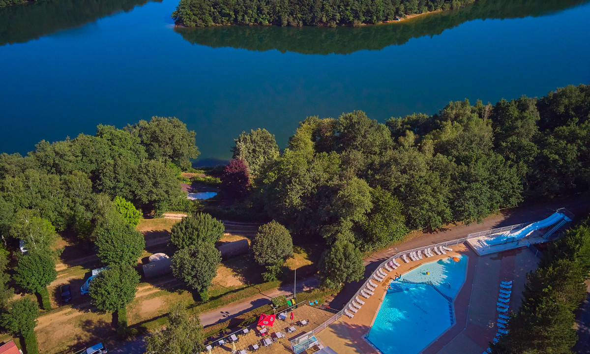 At Terrasses du Lac, an overhead perspective reveals a swimming pool featuring slides adjacent to cabins and trees beside a scenic lake.