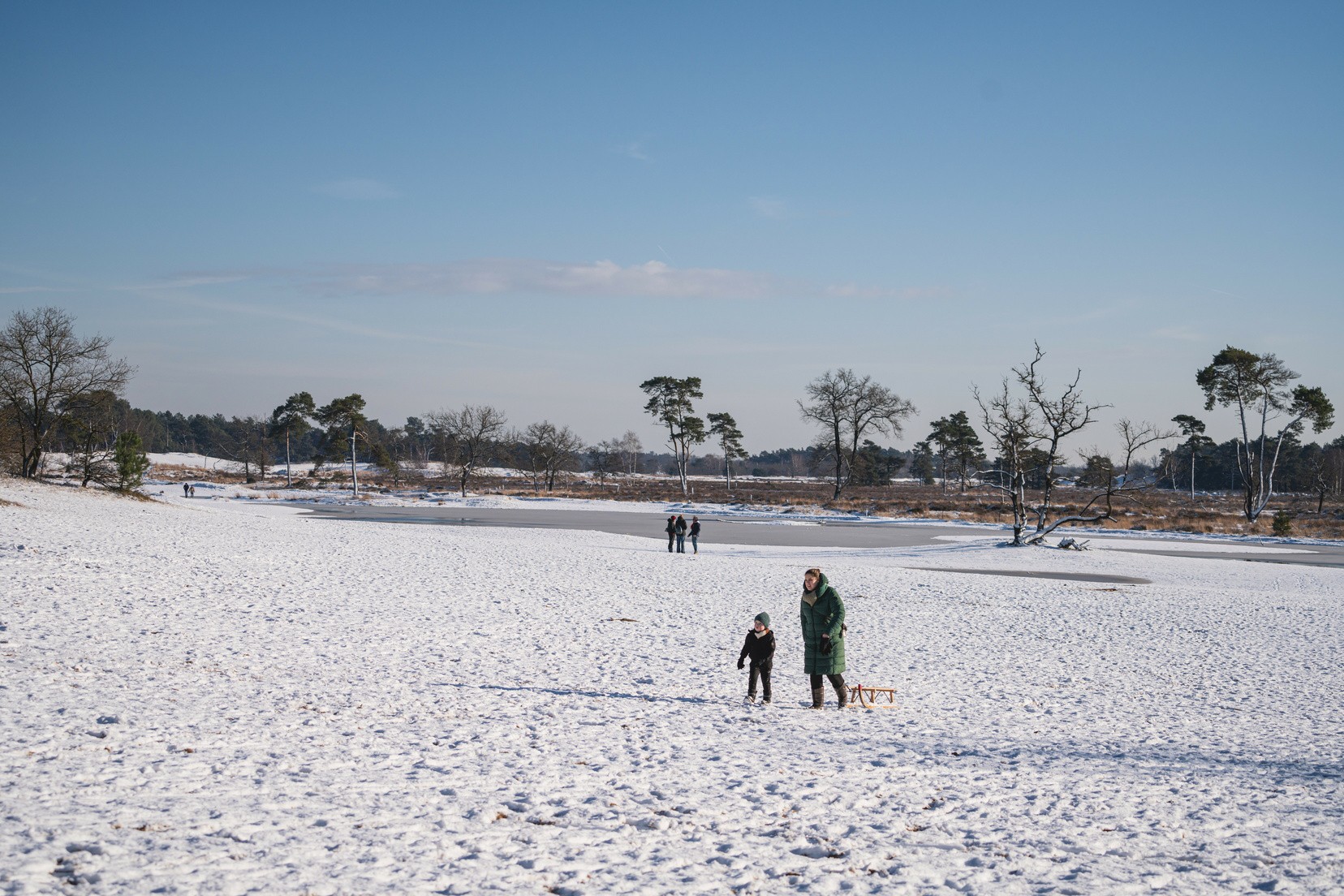People walking across snow-covered field near frozen pond under clear sky; bare trees and distant forest in background at Marvilla Parks Kaatsheuvel
