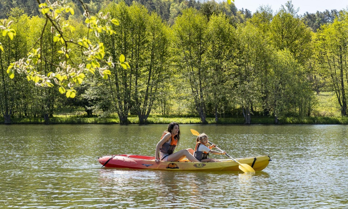 Two people paddle a yellow and red kayak on a serene lake, surrounded by lush, green trees in the background at La Ribeyre