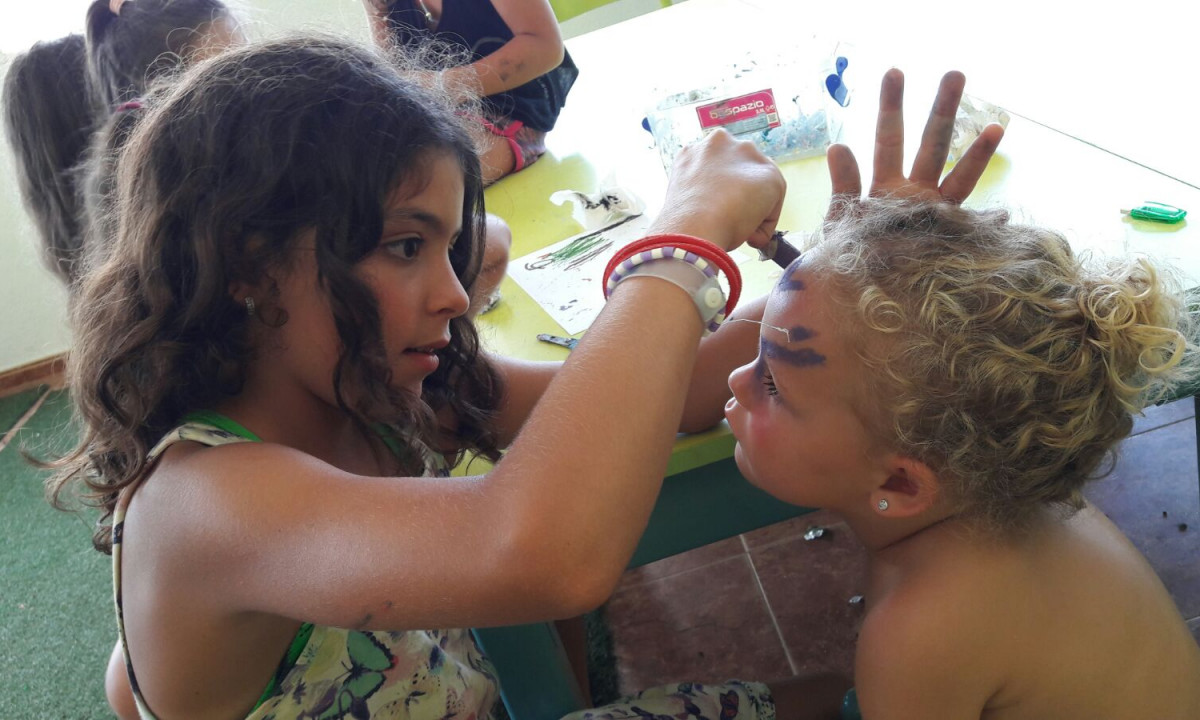 A girl paints another child's face in a brightly lit room, with arts and crafts materials on the table nearby at Resort Els Pins