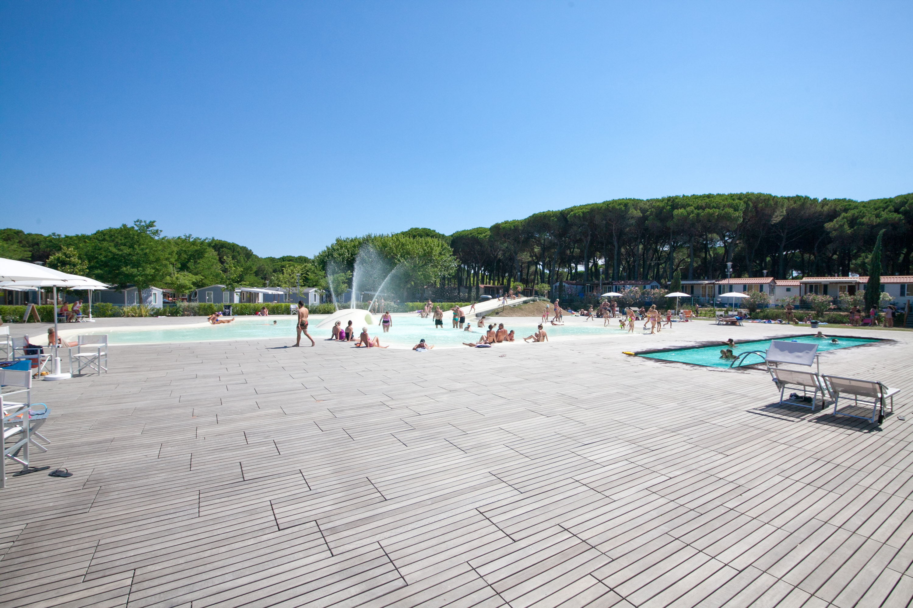 People are enjoying a sunny day, swimming and relaxing by the pool with fountains, surrounded by trees and cabins at Pineta Sul Mare Camping Village