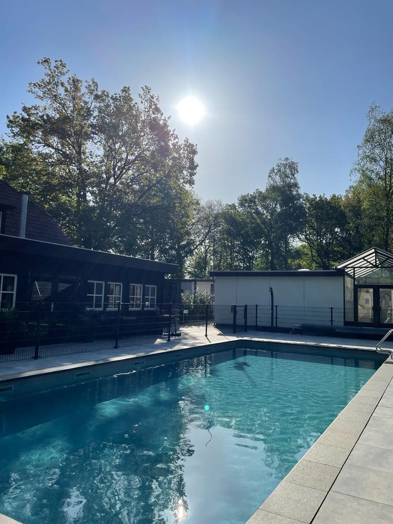At de Vinkenkamp, a swimming pool mirrors sunlight, encircled by trees and structures in a serene, forested environment.
